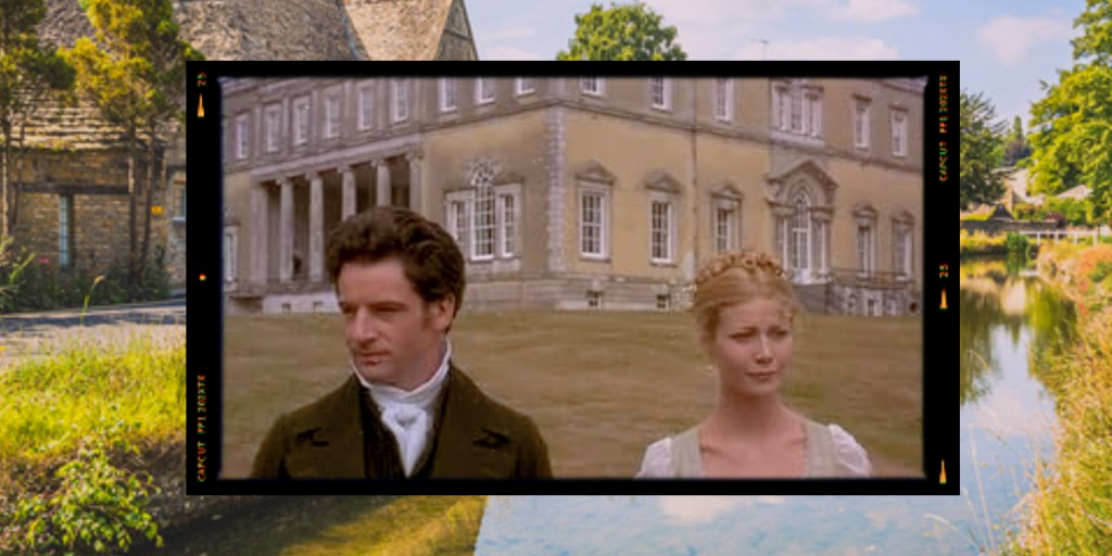 A man and a woman stand outdoors in front of a historical building. The man is wearing a dark coat and the woman is in a light dress with a braided hairstyle. The setting includes greenery and a body of water.