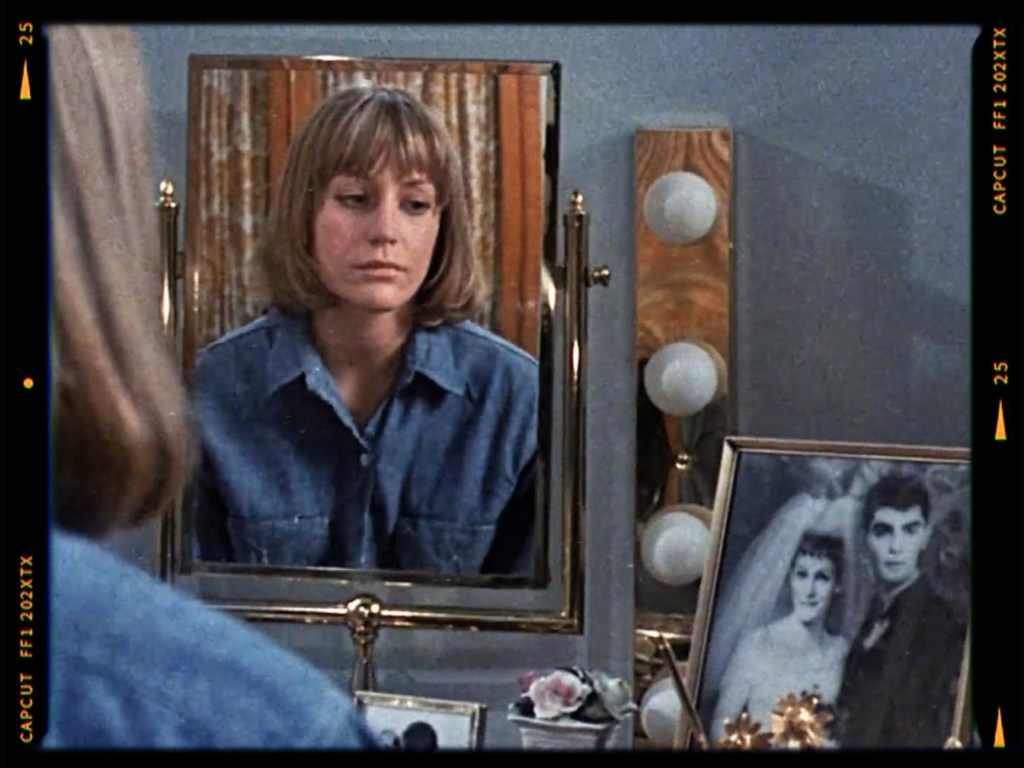 A woman with shoulder-length hair gazes thoughtfully into a mirror, wearing a blue denim shirt. In the foreground, a decorative vanity displays a framed wedding photo and flowers.