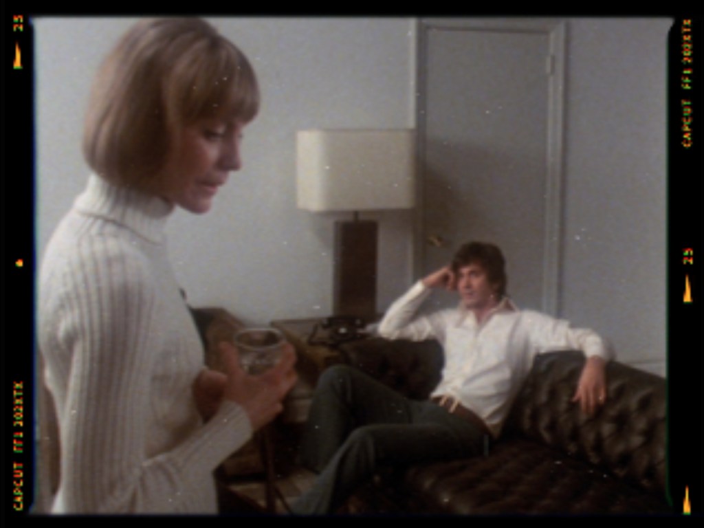 A woman in a white sweater stands holding a glass, looking down, while a man in a white shirt sits on a couch, observing her with a thoughtful expression.