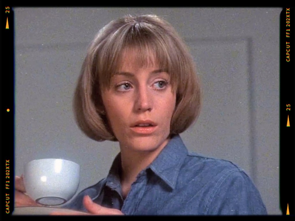 A woman with short blonde hair and a blue shirt, holding a small white cup, looks thoughtfully off-camera.