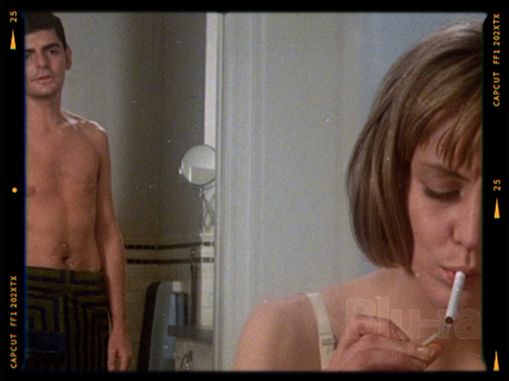 A shirtless man stands in a bathroom while a woman, dressed in a light top, sits nearby smoking a cigarette.