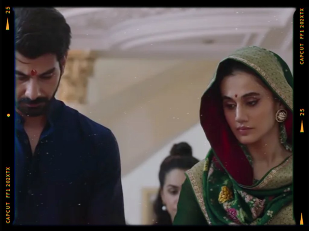 A man and a woman, both dressed in traditional attire, stand side by side indoors. The man wears a dark blue shirt and has a red bindi on his forehead, while the woman wears a green and red embellished outfit with a dupatta. They both appear contemplative, focused on something in front of them.