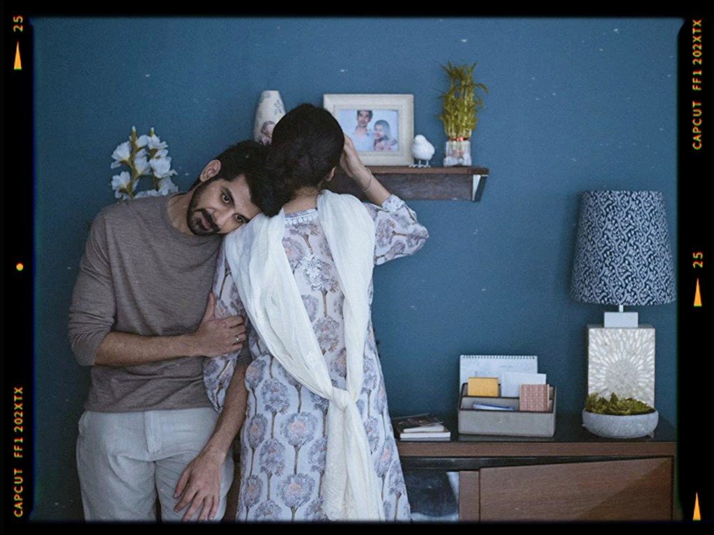 A man leans affectionately on a woman's shoulder in a cozy indoor setting with a blue wall and decorative items on a shelf.