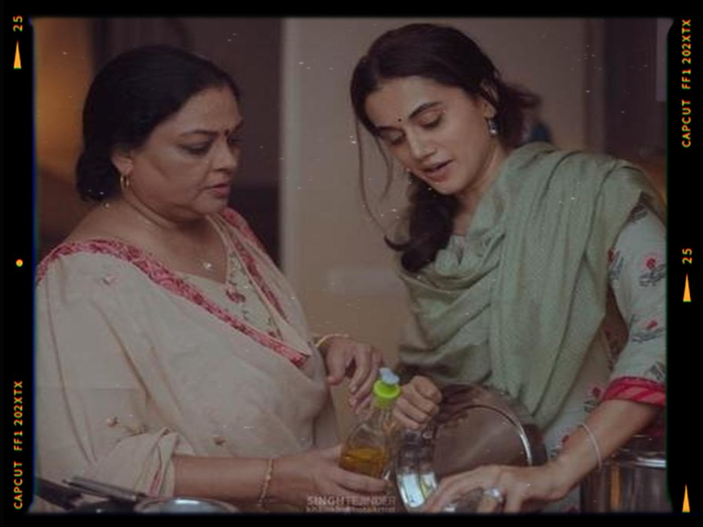 A woman in traditional attire watches as another woman pours cooking oil from a bottle into a pot in a kitchen setting.