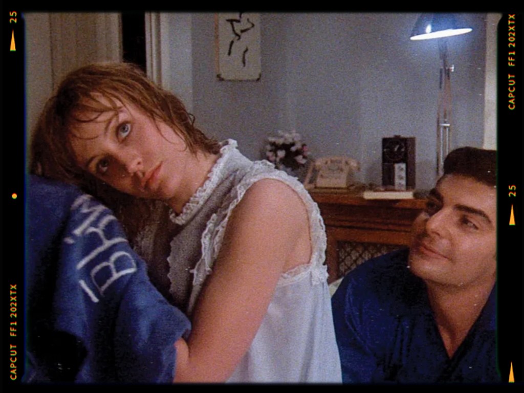 A woman with short hair and a light blue nightgown looks over her shoulder at a man beside her, who is smiling and wearing a dark blue shirt. The background features a softly lit room with a lamp and a clock.