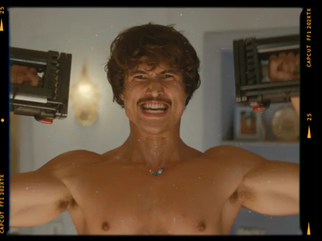 A muscular man smiling broadly while flexing his arms and holding weights in each hand, set in a brightly lit room.