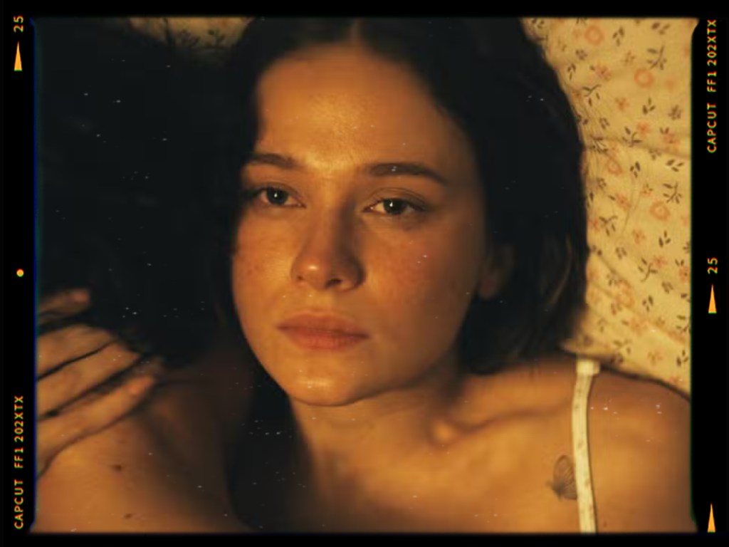 A close-up of a woman with a contemplative expression, lying on a floral-patterned bed, with a hint of her shoulder and another person's arm in the background.