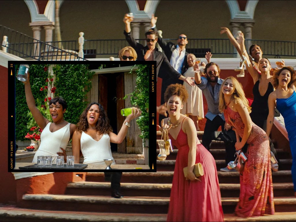 A vibrant group of people celebrating outdoors, smiling and raising their drinks near a beautiful building with red flowers, featuring two women in white dresses in the foreground holding drinks and looking joyful.