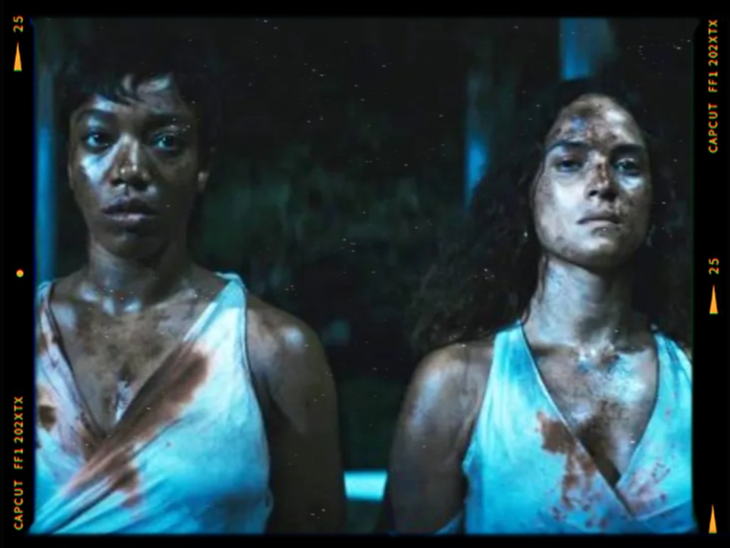 Two women standing side by side, both wearing white dresses, with dirt and stains on their clothes. They appear serious and determined, set against a dark, moody background.