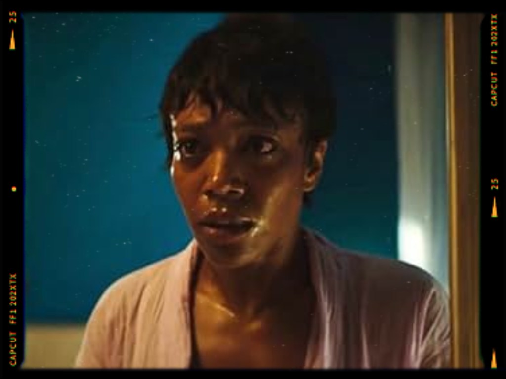 A woman with short hair looks surprised or fearful, wearing a light-colored robe, in a dimly lit room with blue walls.