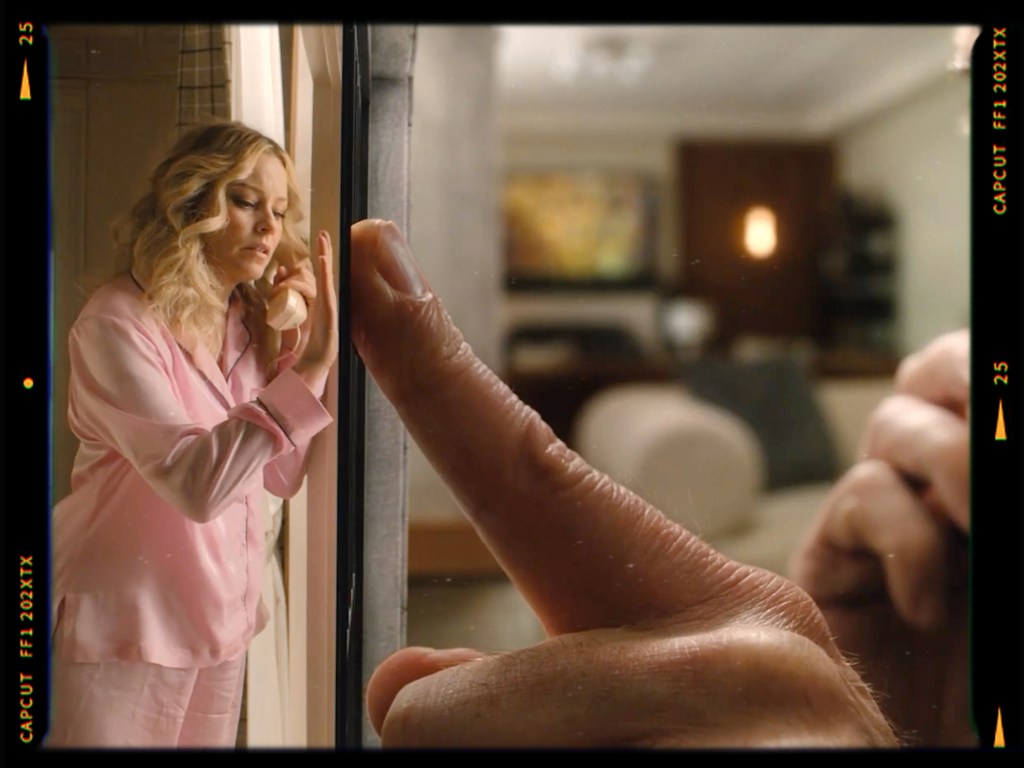 A woman in pink pajamas leans against a glass panel with a thoughtful expression, while a hand reaches toward the glass from the other side.