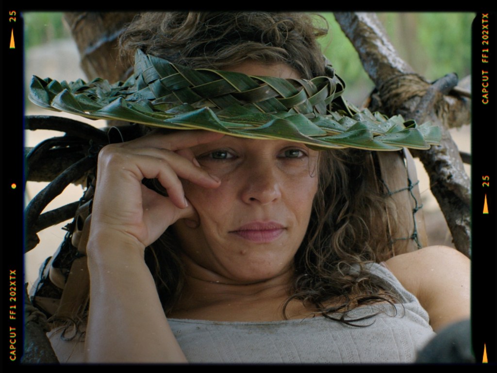 A woman with curly hair wearing a leaf crown, resting thoughtfully while lying on a natural surface.