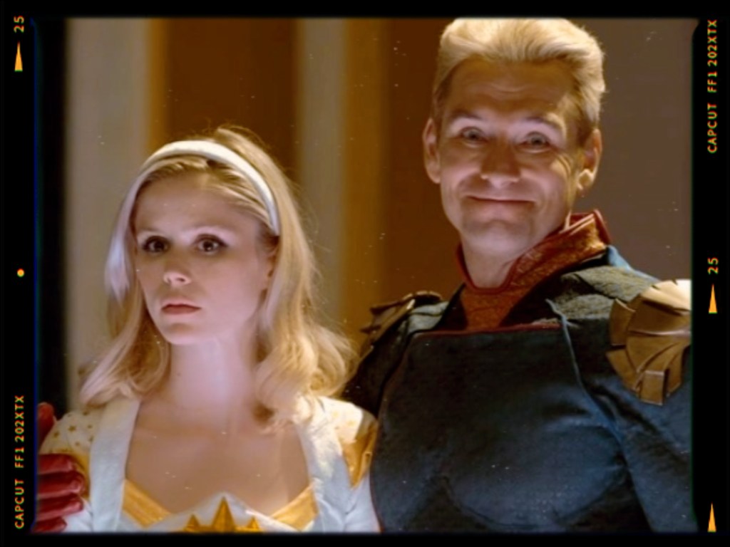 A close-up of two characters, one with blonde hair and a white outfit, and the other with short blonde hair wearing a dark superhero costume with gold accents, both looking serious.