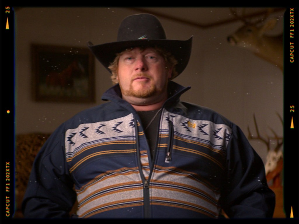 A man wearing a black cowboy hat and a patterned zip-up jacket, with a serious expression, standing in a room with a mounted deer head in the background.