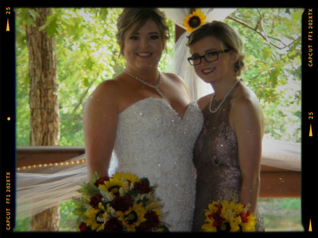 Two women posing together at a wedding celebration, one in a white bridal gown and the other in a sparkly dress, both holding bouquets of flowers.
