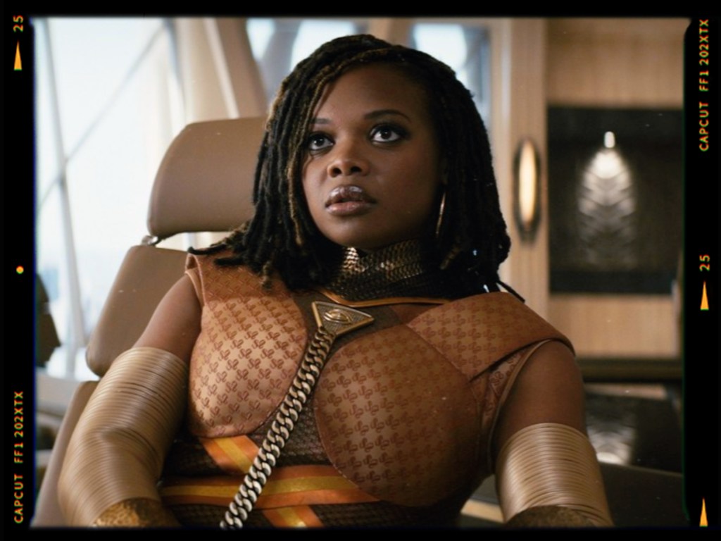 A woman with dreadlocks, wearing a stylish, futuristic outfit, seated in a modern interior setting.