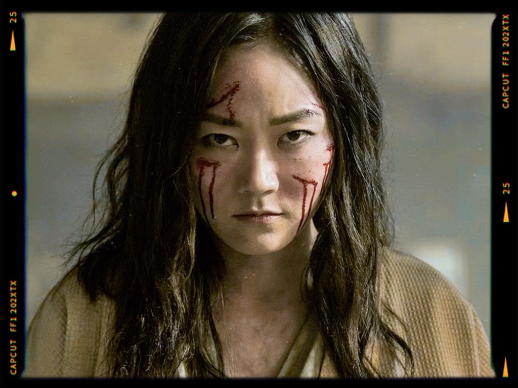 A close-up portrait of a woman with long hair, intense expression, and red streaks on her face, suggesting a dramatic or intense scene.