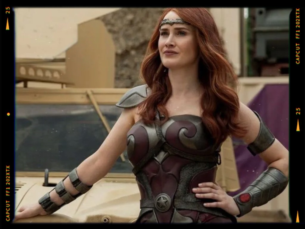 A woman wearing a warrior costume with a breastplate and shoulder armor, standing confidently in front of a military vehicle.