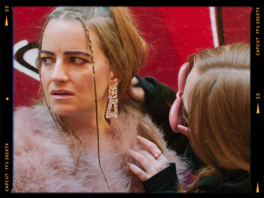 A woman with long hair and dramatic makeup looks surprised while another person styles her hair, set against a vibrant red background. The first woman wears large earrings and a fluffy coat.
