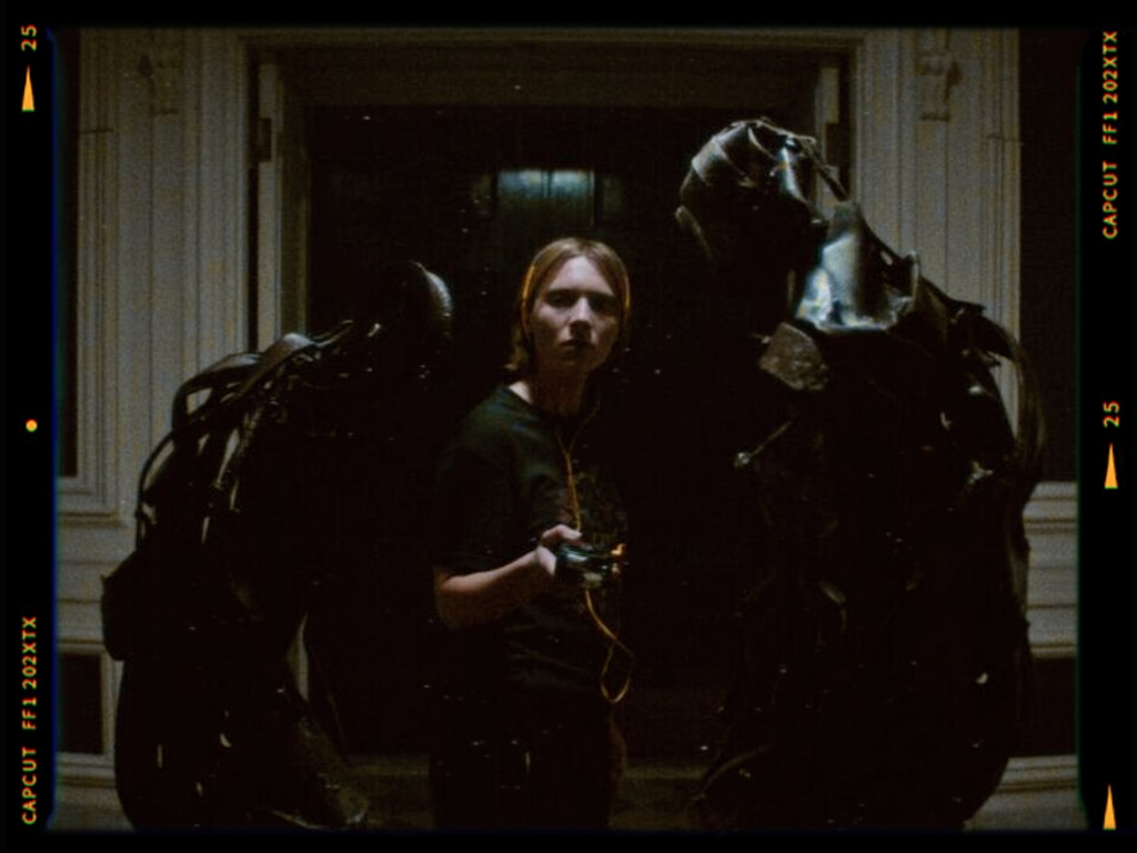 A young person standing in a dimly lit room, holding a device while flanked by two large, dark, humanoid figures.