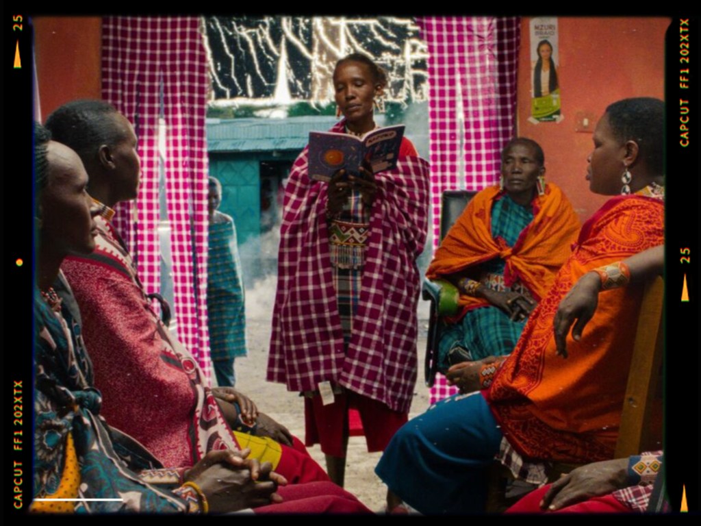A group of women in traditional attire sitting in a room, with one woman reading from a book while others listen attentively.