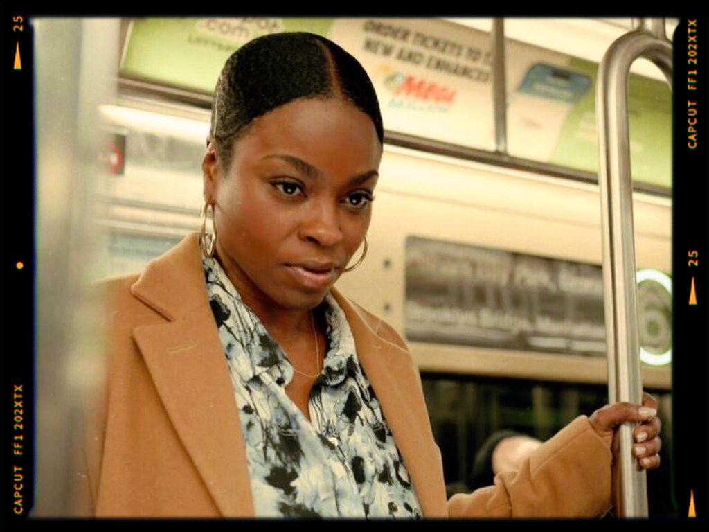 A woman with braided hair wearing a brown coat and patterned blouse standing in a subway train, holding onto a pole and looking pensively.