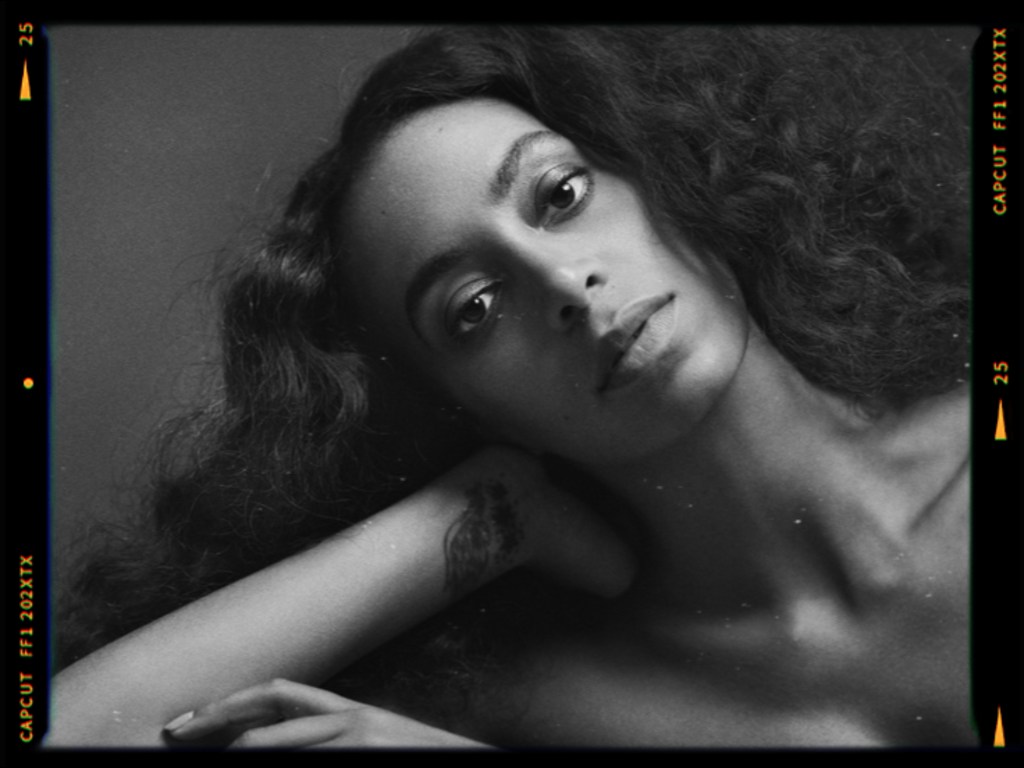 A close-up black and white portrait of a woman with long, curly hair, looking directly at the camera while resting her chin on her hand.