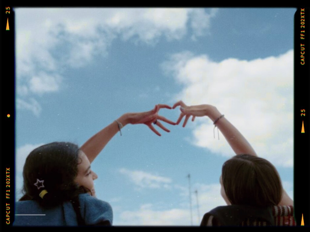 Two people forming a heart shape with their hands against a blue sky with fluffy clouds.