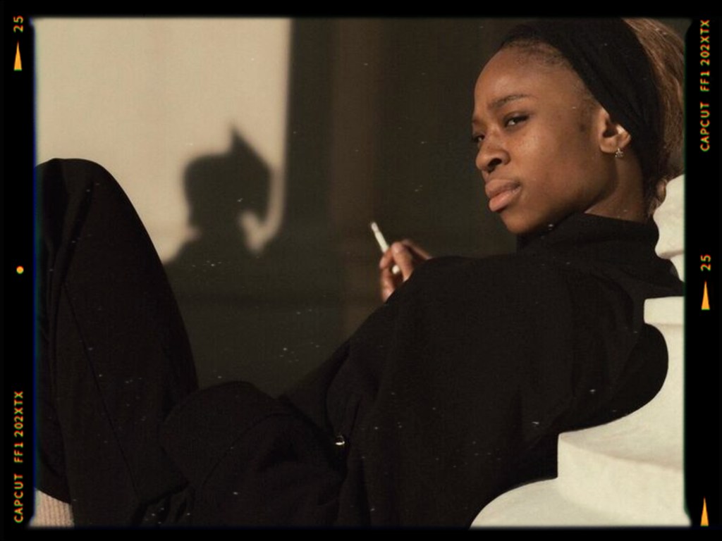 A thoughtful young woman in a black outfit sits against a light background, holding a cigarette, with shadows casting around her.