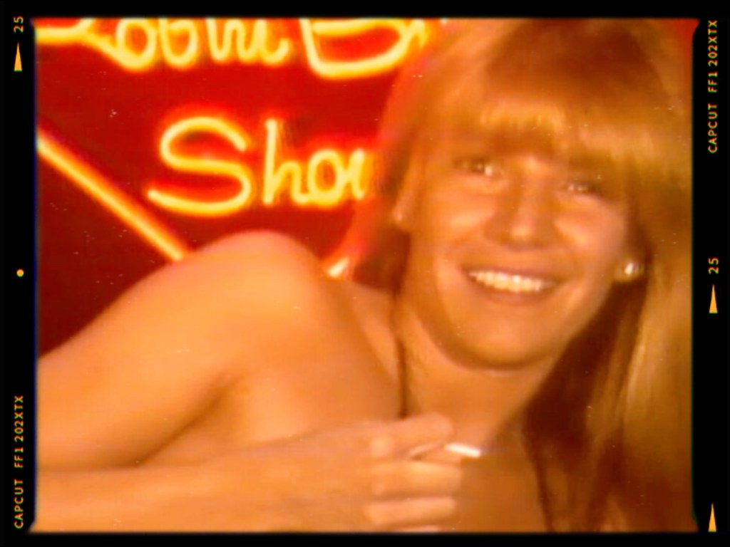 A smiling woman with long hair holding a cigarette, in front of a brightly lit neon sign.
