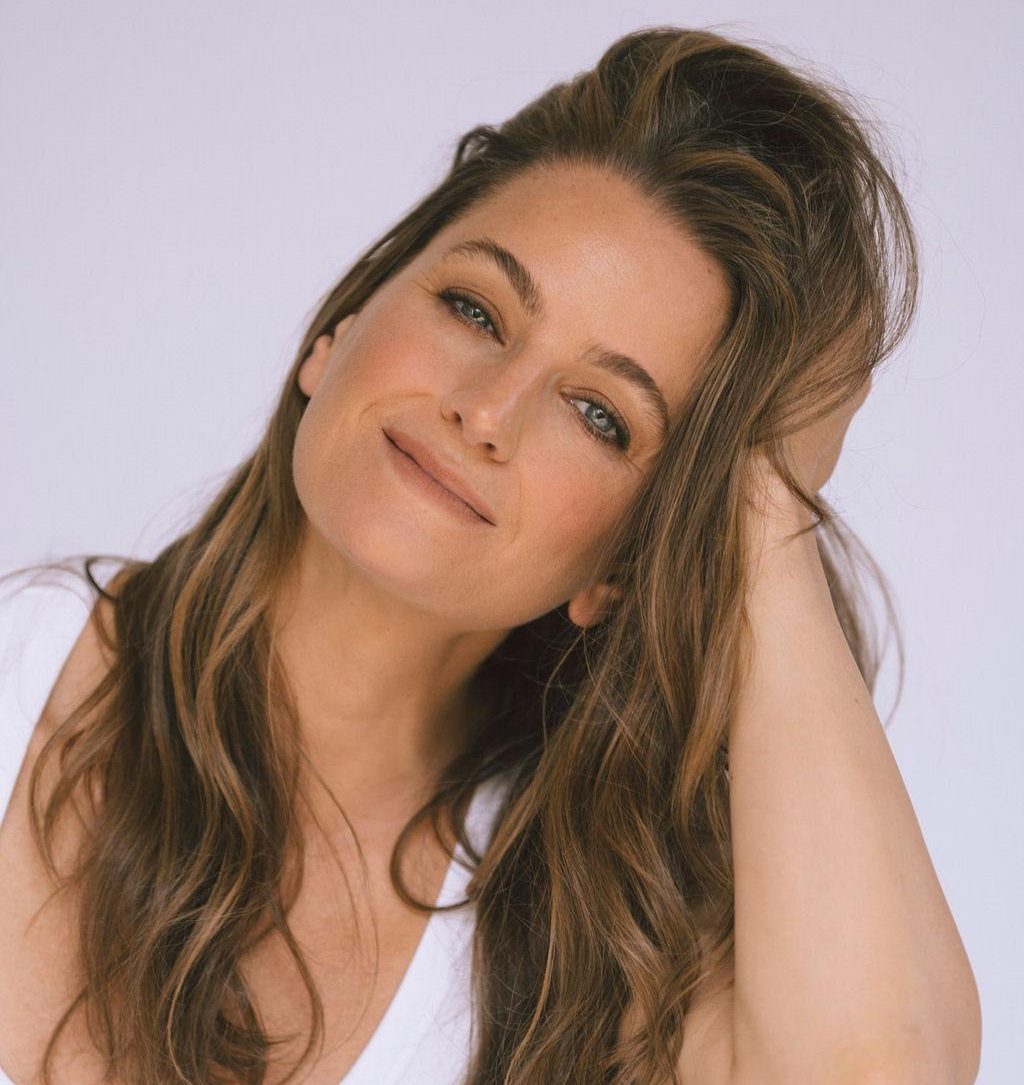 A woman with long brown hair smiles softly while resting her hand on her head, against a light background.